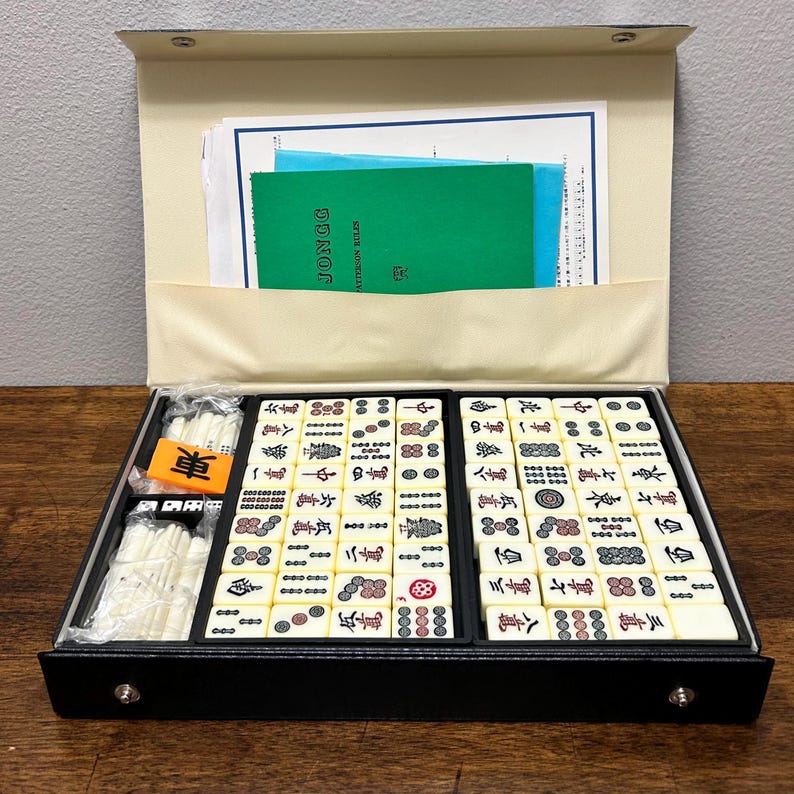May include: A black Mahjong set with ivory tiles, dice, and a wind indicator. The set includes a green booklet labeled "JONGG PATTERSON RULES" and is stored in a black case with a beige interior. The tiles feature Chinese characters and symbols.