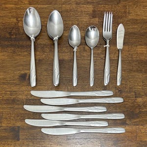 Puede incluir: Una colección de cubiertos de acero inoxidable, que incluye cucharas, tenedores y cuchillos, dispuestos sobre una superficie de madera. La cubertería tiene un patrón floral decorativo en los mangos. El juego incluye varios tamaños de cuchara, un tenedor y cuchillos de mantequilla.