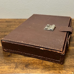 May include: A rectangular brown case with a textured surface and visible stitching along the edges. The case features a silver-toned metal latch mechanism on the top lid and a small matching brown tab closure on the side for securing the lid shut.