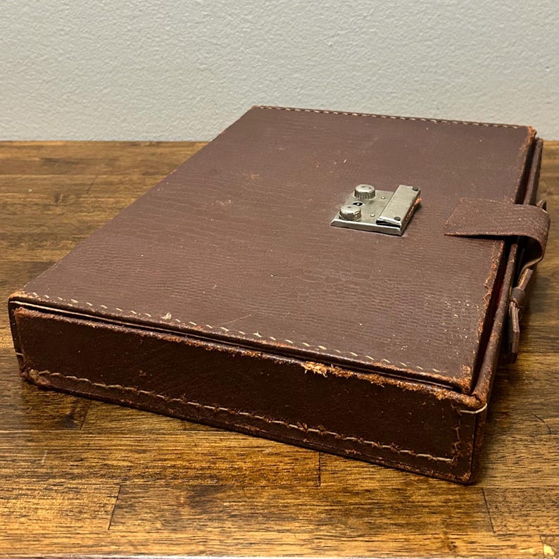 May include: A rectangular brown case with a textured surface and visible stitching along the edges. The case features a silver-toned metal latch mechanism on the top lid and a small matching brown tab closure on the side for securing the lid shut.