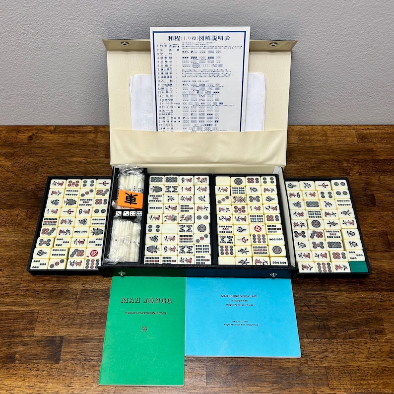 May include: A complete Mahjong set with ivory-colored tiles, dice, and instruction manuals. The tiles are arranged in black trays within a beige case. Two manuals are visible, one green and one blue, with text. The set is displayed on a wooden surface.