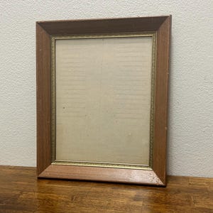 Op de afbeelding: Een rechthoekige fotolijst met een houten buitenrand in een middelbruine tint en een dunne, decoratieve goudkleurige binnenrand. De lijst omsluit een effen, lichtgekleurd passe-partout dat een neutraal vlak biedt voor kunst of foto's.