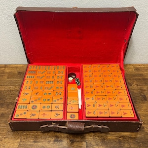 May include: A vintage Mahjong set housed in a brown carrying case with a vibrant red interior lining. The set includes numerous rectangular tiles in a warm tan color, featuring various black, red, green, and blue engraved symbols, characters, and patterns.