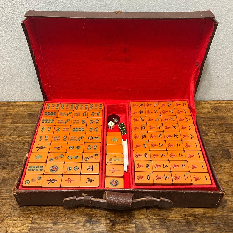 May include: A vintage Mahjong set housed in a brown carrying case with a vibrant red interior lining. The set includes numerous rectangular tiles in a warm tan color, featuring various black, red, green, and blue engraved symbols, characters, and patterns.