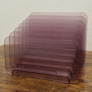 May include: A translucent purple acrylic desk organizer featuring a tiered, stepped design with multiple vertical slots for holding documents, mail, or stationery. The organizer has rounded corners and a smooth, glossy finish throughout its structure.