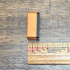 May include: A rectangular block with a smooth orange front face and a black side panel, positioned next to a wooden ruler that shows the object has a width of approximately 0.5 inches.
