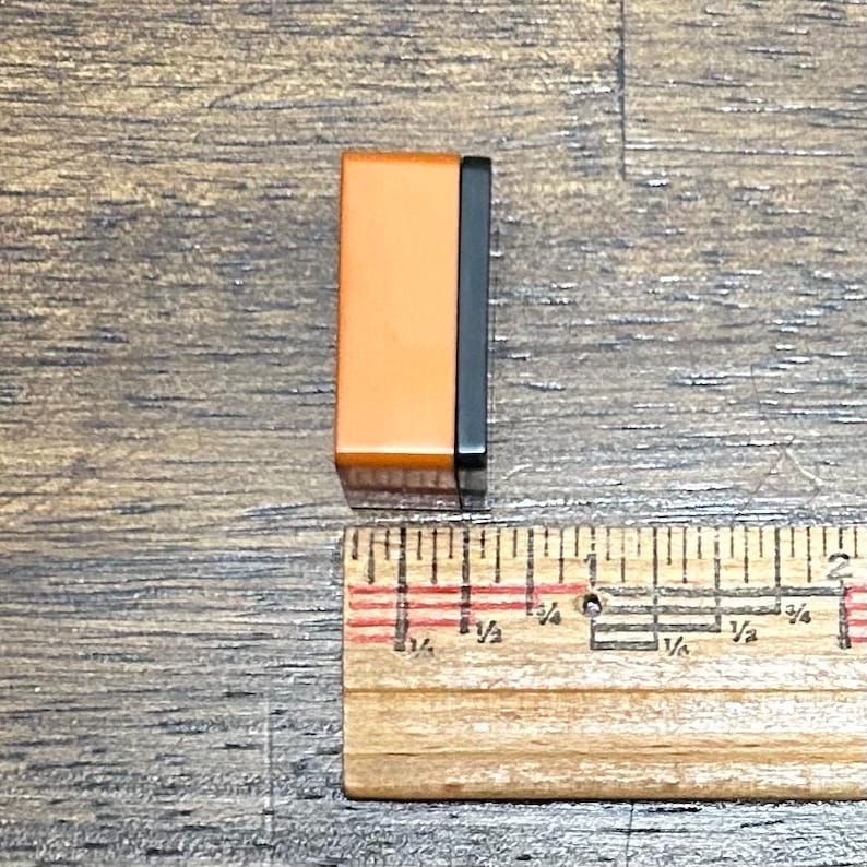 May include: A rectangular block with a smooth orange front face and a black side panel, positioned next to a wooden ruler that shows the object has a width of approximately 0.5 inches.