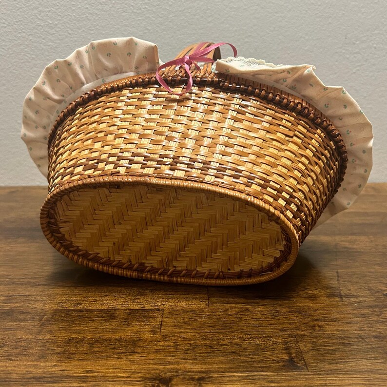 Puede incluir: Una cesta ovalada de mimbre trenzado con un forro de tela clara con un peque&ntilde;o estampado floral verde y un borde con volantes. La cesta incluye un asa de madera marr&oacute;n decorada con una fina cinta rosa atada en un lazo en la parte superior.