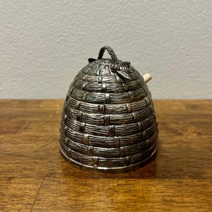 Może przedstawiać: Srebrny pojemnik w kształcie ula o tkanej fakturze. Ma mały uchwyt na górze i drewniany patyczek wystający z boku. Mała ozdoba w kształcie pszczoły jest przymocowana do góry.