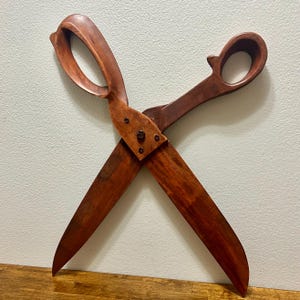 May include: A large, decorative pair of scissors carved from dark brown wood with a visible grain. The scissors feature two rounded handles and long, pointed blades connected at the center by a dark metal bolt and three smaller metal screws.
