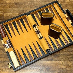 Puede incluir: Juego de backgammon abierto con marco negro, superficie de juego beige y puntas triangulares marrones y negras. Incluye cubiletes marrones, dados y fichas. El juego se muestra sobre una superficie de madera.