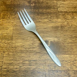 May include: A stainless steel fork with four tines. The handle features a decorative design with small star-like shapes. The fork is set against a wooden surface.