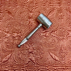 May include: A silver-colored gavel with a cylindrical head and a long, slender handle. The gavel is resting on a textured, reddish-orange fabric with an intricate, quilted pattern. The gavel is a tool often used in legal or auction settings.
