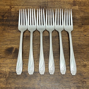 May include: A set of six silver-toned metal forks arranged in a row. Each fork features four tines and a handle decorated with a raised floral motif at the base. The metal has a smooth, matte finish, and the design is consistent across all six pieces.