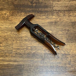 May include: A dark brown corkscrew with a lever design. The corkscrew has a metal worm and a handle with a curved design. The lever arms are a lighter brown colour. The corkscrew is on a wooden surface.