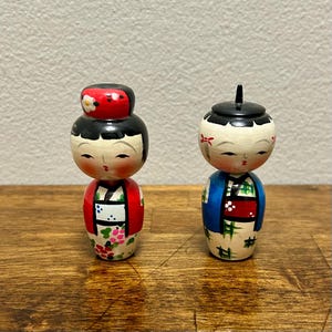 May include: Two Japanese Kokeshi dolls. The dolls have painted faces, black hair, and wear traditional clothing. One doll has a red hat with a white flower, and the other has a black hat. The dolls are on a wooden surface.