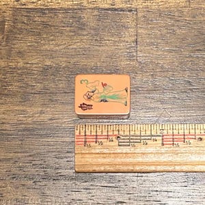 May include: A rectangular mahjong tile with a tan surface featuring a green number 3, a black Chinese character, and a colorful illustration of a person in traditional robes. A wooden ruler is positioned below, showing a width of approximately 1 inch.