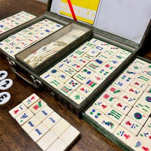May include: A vintage Mahjong set with ivory-colored tiles, featuring various symbols and characters in red, green, blue, and black. The tiles are arranged in a green patterned case with multiple compartments. Additional white and black game pieces are visible.