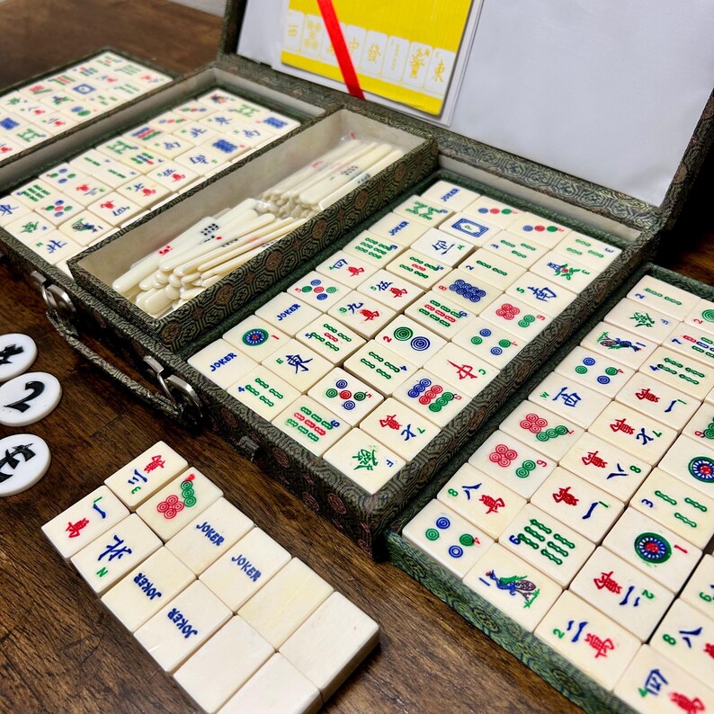 May include: A vintage Mahjong set with ivory-colored tiles, featuring various symbols and characters in red, green, blue, and black. The tiles are arranged in a green patterned case with multiple compartments. Additional white and black game pieces are visible.