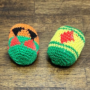 May include: Two small, round, crocheted hacky sacks. The left one is orange with black and red triangular patterns and a green base. The right one is yellow with a red diamond pattern and green bands at the top and bottom, featuring a green base.