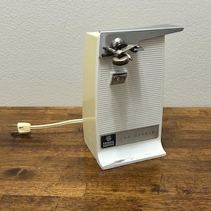 以下が含まれることがあります： ヴィンテージのゼネラル・エレクトリック製電動缶切り。クリーム色の本体に白いフロントパネル、シルバーの金属製オープナーが付いています。「CAN OPENER」の文字が前面に印刷されています。2ピンのプラグが付いた電源コードがベースから伸びています。