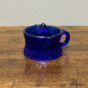 May include: Cobalt blue glass citrus juicer with a spiral design on the exterior. The juicer has a conical reamer in the center and a small handle. The juicer is sitting on a wooden surface.