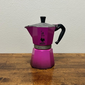 May include: A magenta Moka Color stovetop espresso maker with a black handle and lid knob. The coffee pot has the Bialetti logo and text on the front. The espresso maker sits on a wooden surface.