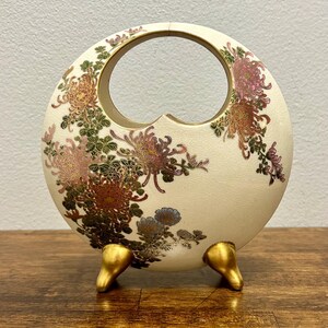 以下が含まれることがあります： 中央に切り抜かれたハンドルと3つの金色の脚を持つ、円形のオフホワイトの陶器製花瓶。表面には、ピンク、オレンジ、青の色合いの菊の花と緑の葉、そして金色の輪郭が描かれた詳細な花柄が施されています。