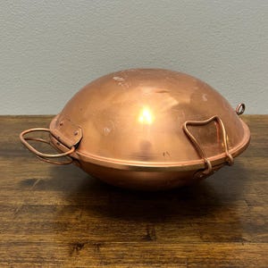 May include: A copper-colored, domed cooking pot with a lid and two handles. The pot has a rounded base and a lid that fits snugly over the top. The handles are made of the same copper material and are attached to the sides of the pot.