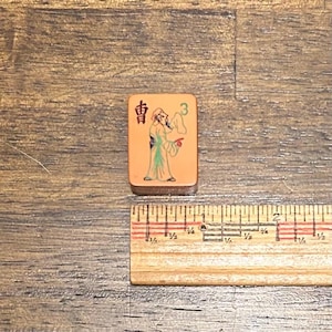 May include: A rectangular tan mahjong tile featuring a black Chinese character, a green number 3, and a colorful illustration of a person in traditional robes. A wooden ruler placed below shows the tile is approximately 1.25 inches in height.