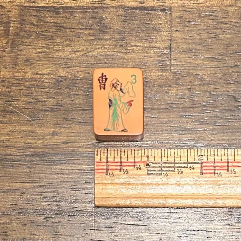 May include: A rectangular tan mahjong tile featuring a black Chinese character, a green number 3, and a colorful illustration of a person in traditional robes. A wooden ruler placed below shows the tile is approximately 1.25 inches in height.