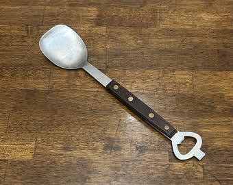 May include: A silver-toned metal spoon with a square-shaped bowl and a dark brown handle secured by three brass rivets. The handle extends into a metal bottle opener at the end, featuring a cutout design for opening beverage bottles.