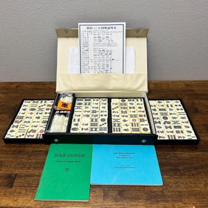 May include: A vintage Mahjong set with ivory-colored tiles, black trays, and a beige case. The set includes dice, tile racks, and instruction booklets. The text "MAH JONGG" is visible on a green booklet.