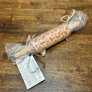 May include: A light brown wooden rolling pin featuring an embossed cherry pattern across its surface. The rolling pin is wrapped in clear plastic packaging tied with twine and includes a white rectangular product tag with printed black text.