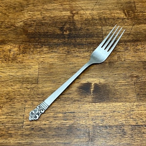 May include: A stainless steel fork with four tines. The handle has a decorative, ornate design at the end. The fork is set against a dark wood grain background.
