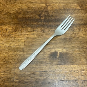 May include: A stainless steel fork with four tines and a decorative floral pattern on the handle. The fork is set against a wooden background.