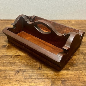 May include: A rectangular wooden caddy with a dark brown stained finish, featuring a central arched handle and decorative scalloped side panels. The open-top container has a flat base and visible wood grain patterns throughout the structure.