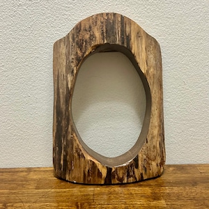 May include: A vertical, rectangular wooden frame with a rounded top arch and a large, central oval cutout. The wood features a natural, raw finish with visible dark brown grain patterns, knots, and textured edges throughout the entire structure.