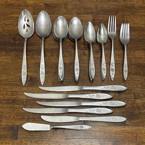 Pode incluir: Uma coleção de talheres vintage, incluindo colheres, garfos e facas, dispostos sobre uma superfície de madeira. Os talheres têm um desenho floral nas alças. As colheres incluem colheres de servir, colheres de sopa e colheres de chá. As facas são facas de manteiga e facas de jantar.