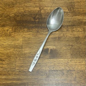 Peut inclure: Une cuillère en argent avec un long manche et un bol ovale. Le manche est orné d'un motif floral. La cuillère est posée sur un fond en bois.