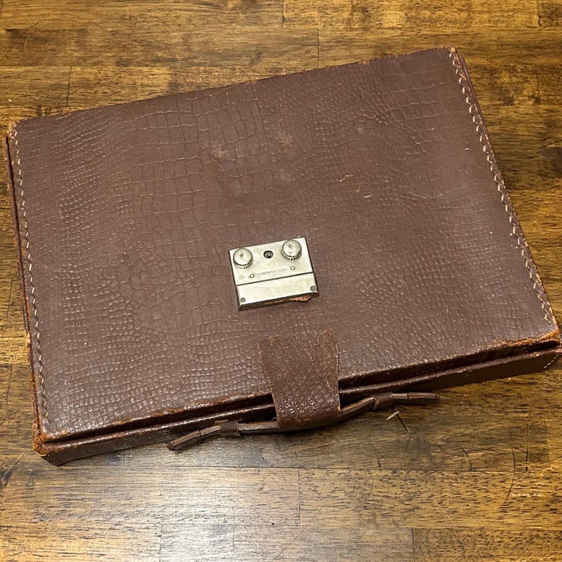 May include: A rectangular brown case with a textured crocodile-patterned surface. The case features a central silver-toned metal locking mechanism with two circular knobs and a small tab closure at the bottom edge, secured by a leather strap.