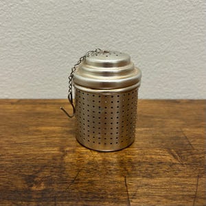 May include: A silver tea infuser with a perforated cylindrical body and a domed lid. A small chain is attached to the lid, ending in a hook. The infuser is designed for steeping loose leaf tea.