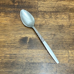 May include: A silver-toned metal spoon featuring a smooth, oval-shaped bowl and a slender handle. The upper portion of the handle is decorated with a detailed, repeating floral pattern, while the lower section remains plain and flat.