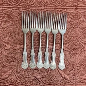 May include: A set of six silver-toned metal forks arranged in a row. Each fork features a four-tine head and a handle decorated with an embossed floral pattern at the base. The metal has a polished finish with visible detailing on the handles.