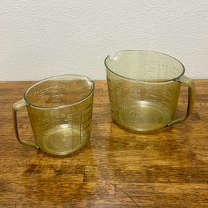 May include: Two clear, angled measuring cups with handles. The cups are made of a translucent, yellowish-green plastic and feature black measurement markings in cups, fluid ounces, and tablespoons. They sit on a wooden surface.