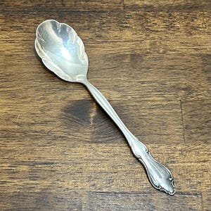 May include: A silver-toned metal spoon featuring a scalloped bowl edge and a decorative handle with embossed detailing. The spoon has a polished finish and a slender neck connecting the bowl to the ornate, flared end of the handle.