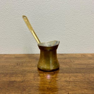 Puede incluir: Una cafetera de latón vintage con un mango largo y un pequeño pico. La olla tiene una base redondeada y una parte superior ligeramente ensanchada. El mango tiene un pequeño agujero en el extremo. La olla está sobre una superficie de madera.