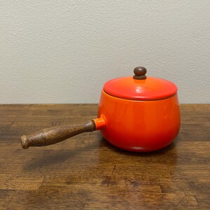 May include: A bright orange enameled cast iron fondue pot with a matching lid and a dark brown wooden handle. The pot features a smooth, glossy finish that transitions from a vibrant orange to a lighter, yellowish-orange hue on the lid.