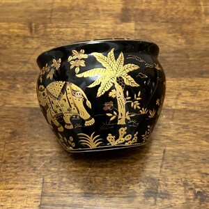 May include: A small, rounded black ceramic bowl featuring intricate gold-colored illustrations. The design depicts a walking elephant, a large palm tree, and various floral and desert-like plant motifs encircling the exterior surface.