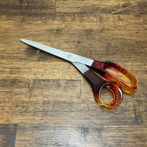 Puede incluir: Tijeras con hojas plateadas y mangos translúcidos rojos y naranjas. Las tijeras están abiertas, mostrando las hojas afiladas. Los mangos tienen un diseño redondeado. Las hojas miden unos 20 cm.